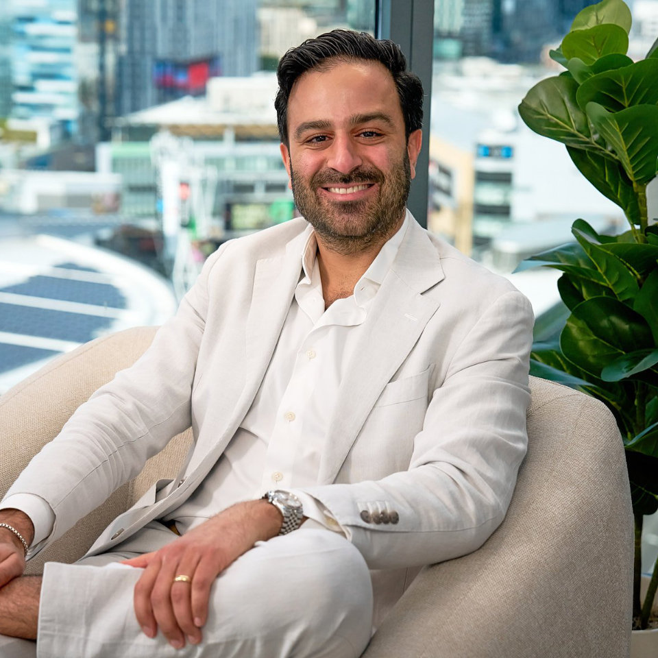 A man in a light-colored suit sits on a beige chair in an office with large windows and city buildings in the background. A plant is visible to the right.