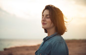 Great pleasure. Charming calm young woman is standing near sea with closed eyes and expressing delight. She is posing against wonderful sunset while enjoying last rays of the sun