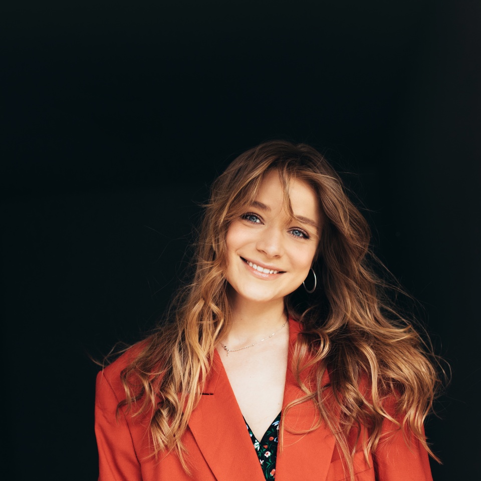 Young beautiful cheerful woman looks straight and poses on camera. Girl in orange jacket stand alone in studio and smiles. Isolated over dark background