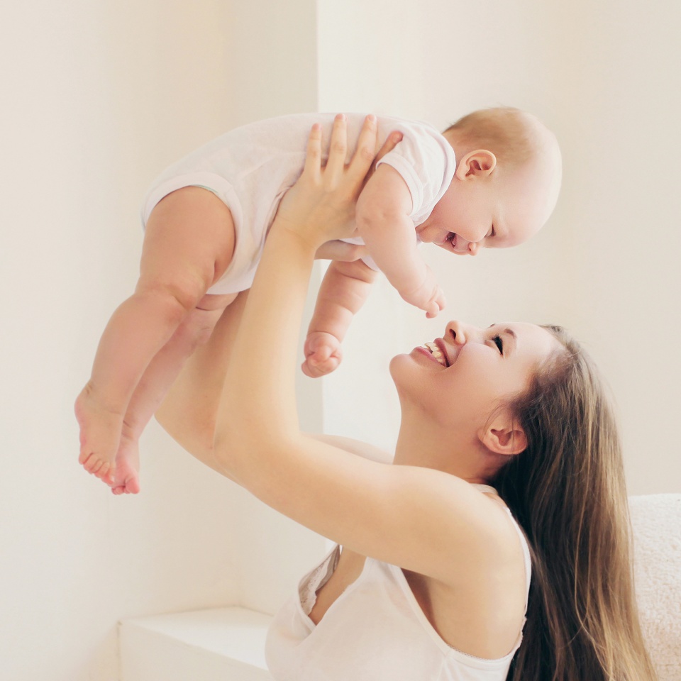 Happy smiling mother and little baby sitting together in white room at home