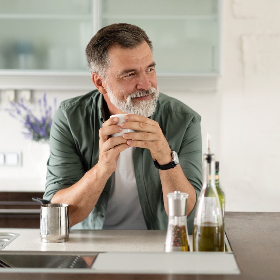 Happy mature man drinking coffee at home in the kitchen, enjoying hot drink in the morning on weekend