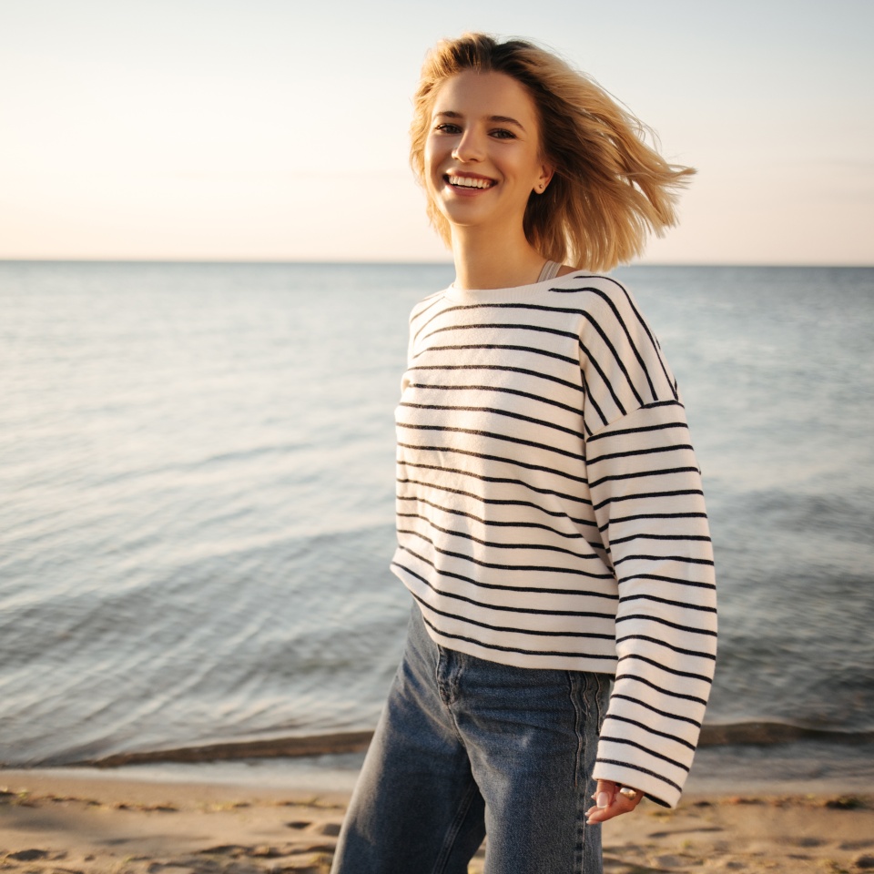 Cheerful beautiful caucasian young woman smiling with teeth looking into camera on background of sea. Her blonde short hair blowing in wind, wearing striped blouse and jeans.