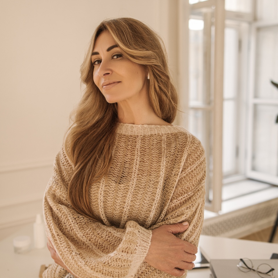 Adult caucasian woman with sly look looks at camera while standing in cozy light living room. Lady with long blond hair is wearing sweater. Leisure lifestyle and beauty concept.
