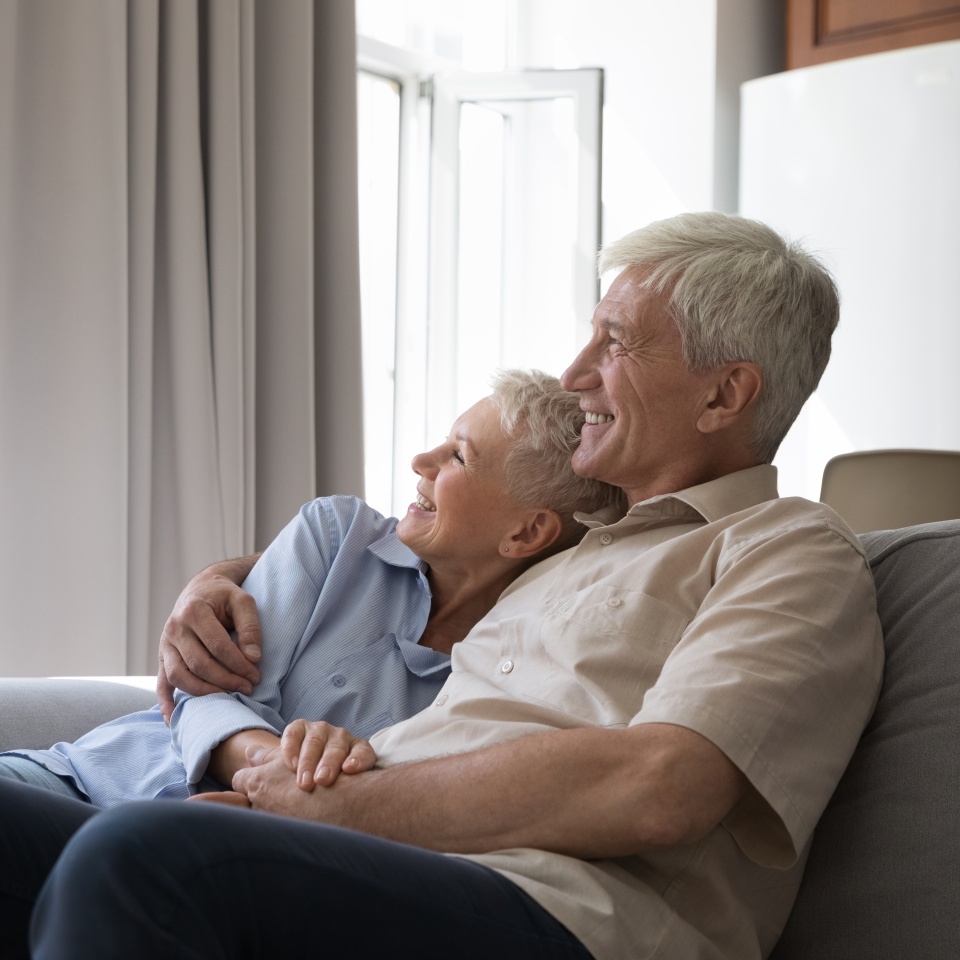 Laughing older grey-haired couple in love hugging daydreaming look out window relax on cozy sofa enjoy pleasant talk spend free time at modern home. Happy marriage, harmony, romantic relations concept