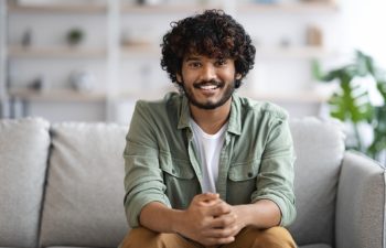 Cheerful millennial curly bearded indian guy in casual outfit sitting on couch at home, holding hands in lock and smiling at camera, young eastern man posing in living room, copy space