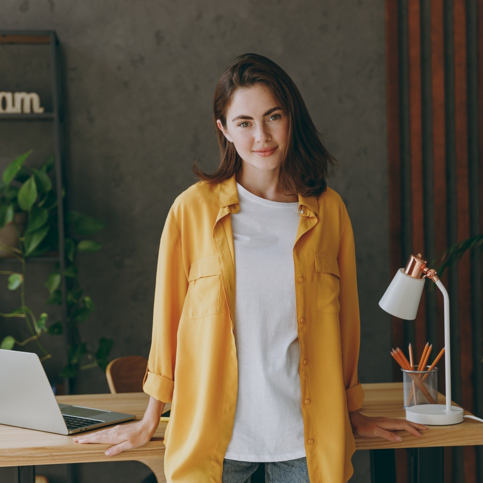 Young smiling happy fun calm happy european successful employee business woman 20s she wear casual yellow shirt stand look camera work at wooden office desk with pc laptop. Achievement career concept