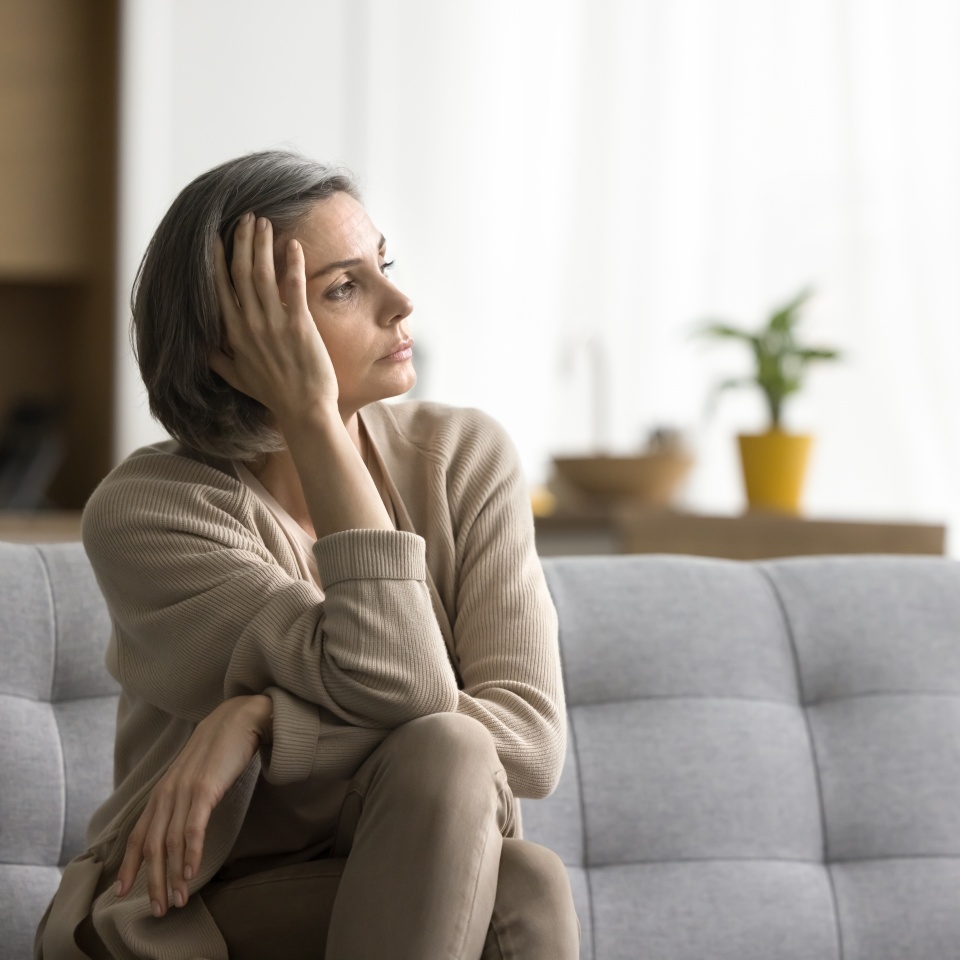 Sad thoughtful grey haired woman sitting on sofa at home, looking at window away in deep unhappy thoughts, leaning head on hand, touching face, suffering from depression, apathy