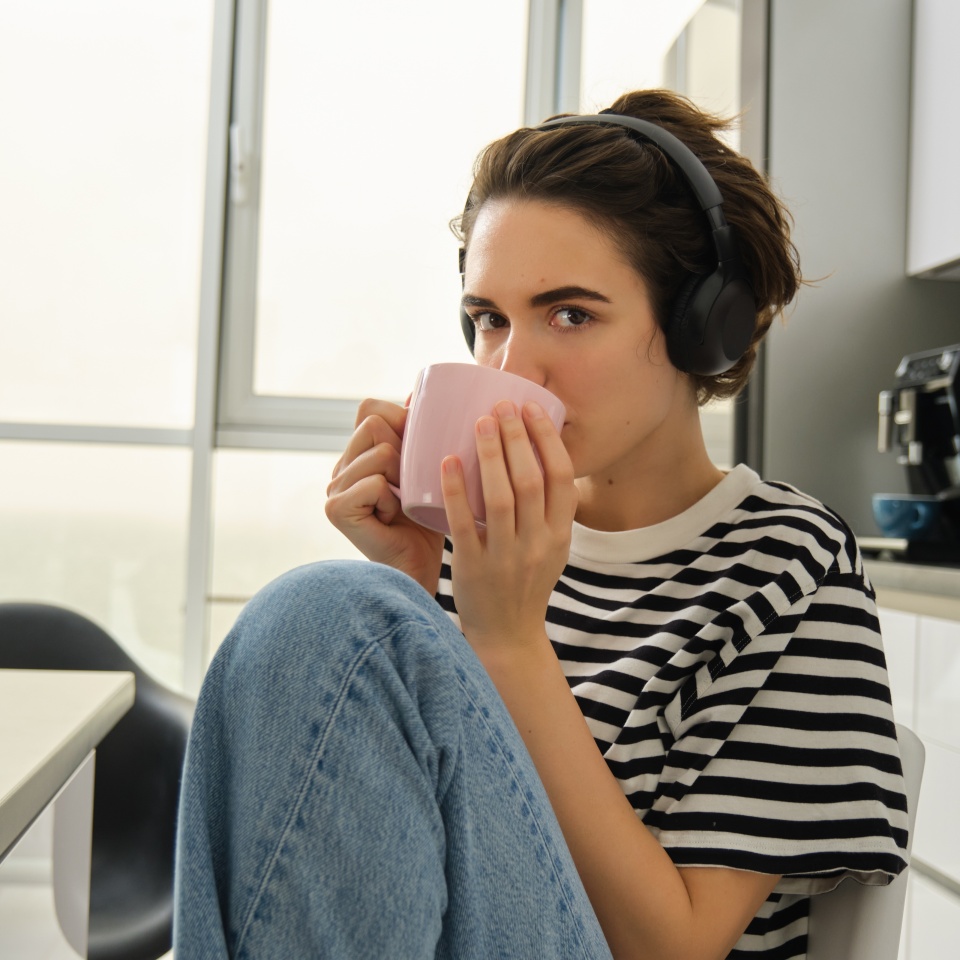 Close up portrait of smiling brunette woman, student drinks her tea and listens music or ebook in headphones, sits in kitchen and relaxes.