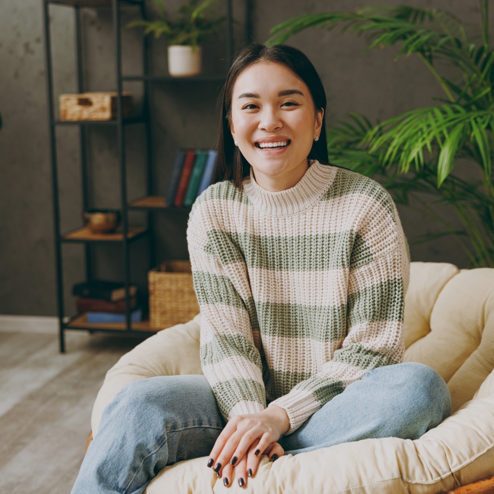 Young smiling happy woman of Asian ethnicity wear casual clothes looking camera sits on armchair stay at home hotel flat rest relax spend free spare time in living room indoor. People lounge concept