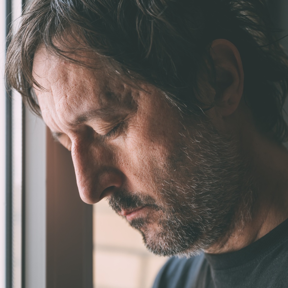 Downhearted man with eyes closed leaning his head close to the apartment window. Depressed unhappy male sad and alone at home. Selective focus.