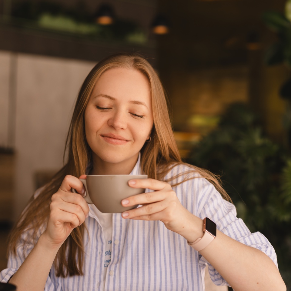 Portrait of gorgeous blonde smiling lady smelling, enjoying of coffee and drinking cappuccino from cup while resting in restaurant. Woman wear stripped shirt.