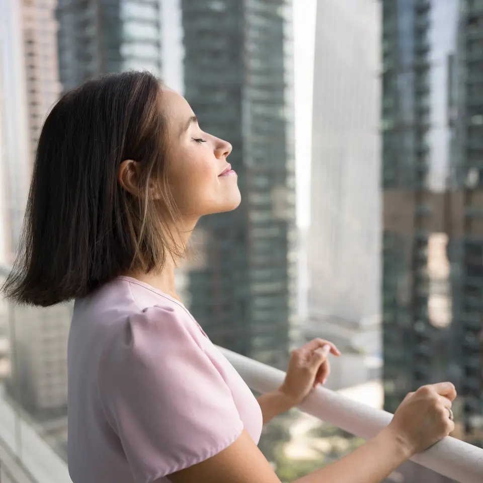 Woman with short dark hair stands on a balcony, eyes closed, facing sunlight, with tall buildings in the background.