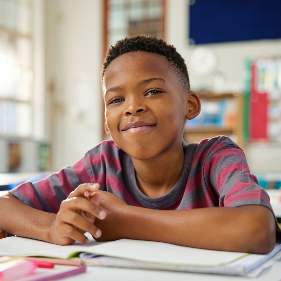 Portrait of happy black elementary boy studying in classroom while looking at camera. Close up face of little satisfied black boy writing in notebook using pencil. Proud schoolchild in primary school.