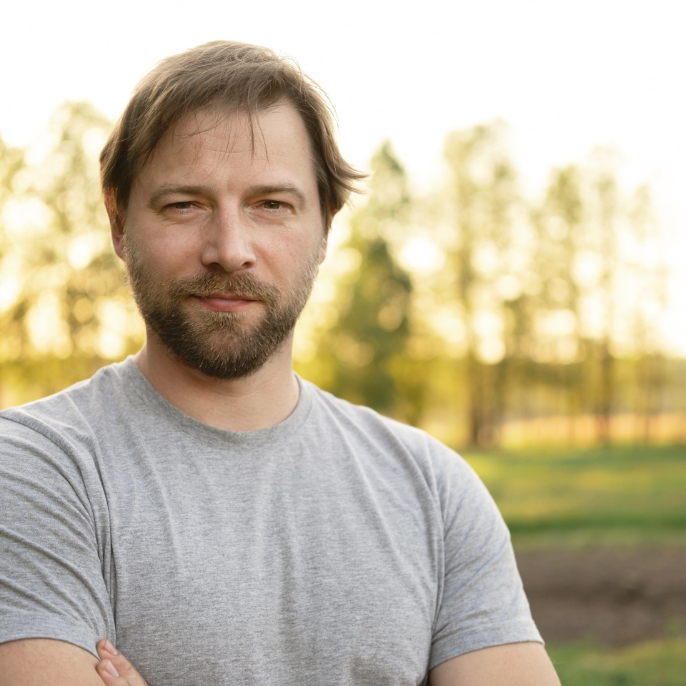 Portrait of handsome young man in gray t-shirt outdoors. Male with long hair and beard feel happy, calm and serious on Green nature sunset background. Man looking away into distance. Diversity People