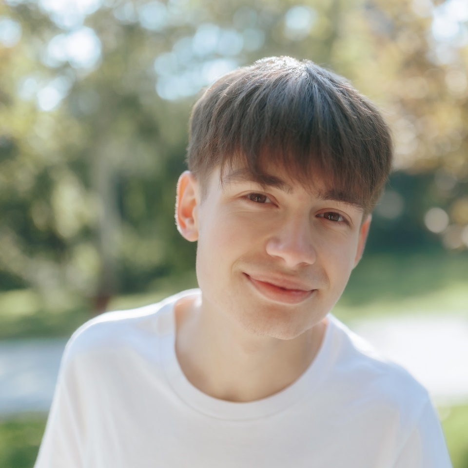 A cheerful adolescent boy, dressed in a simple white shirt, enjoys the outdoors. The sunny park surroundings provide a fresh, natural backdrop for the portrait.