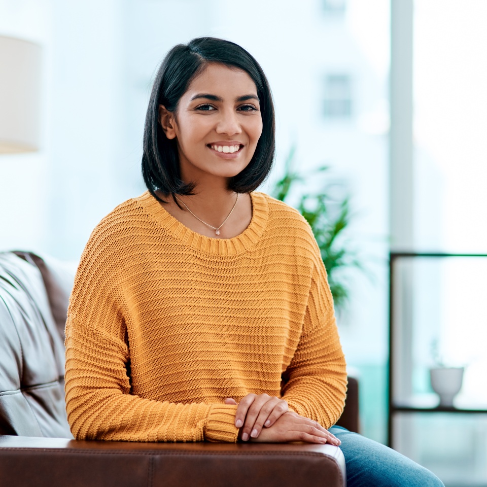 Portrait, relax and smile of Indian woman on sofa in living room of home for weekend break or rest. Calm, easy and peace with happy person in apartment for morning stress relief, time off or wellness