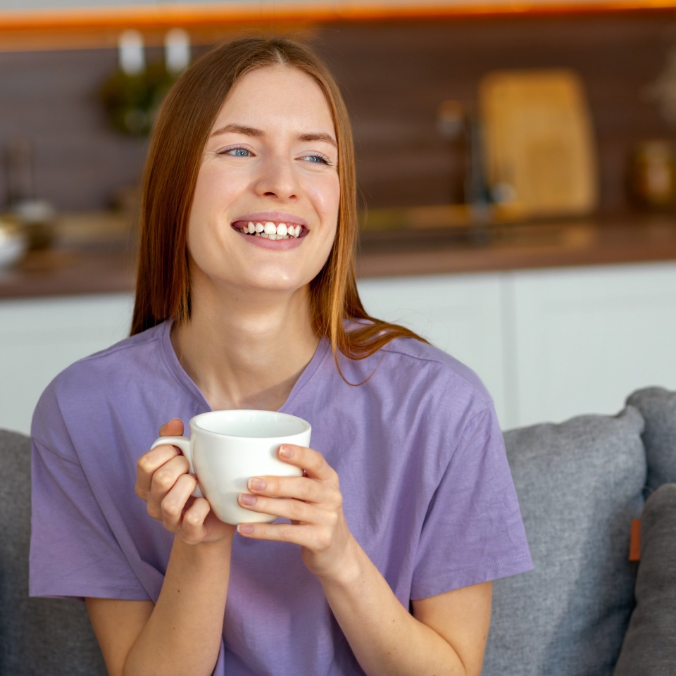Happy young woman holding a coffee mug, enjoying her morning at home with a bright smile, sitting comfortably on a sofa in a cozy kitchen setting