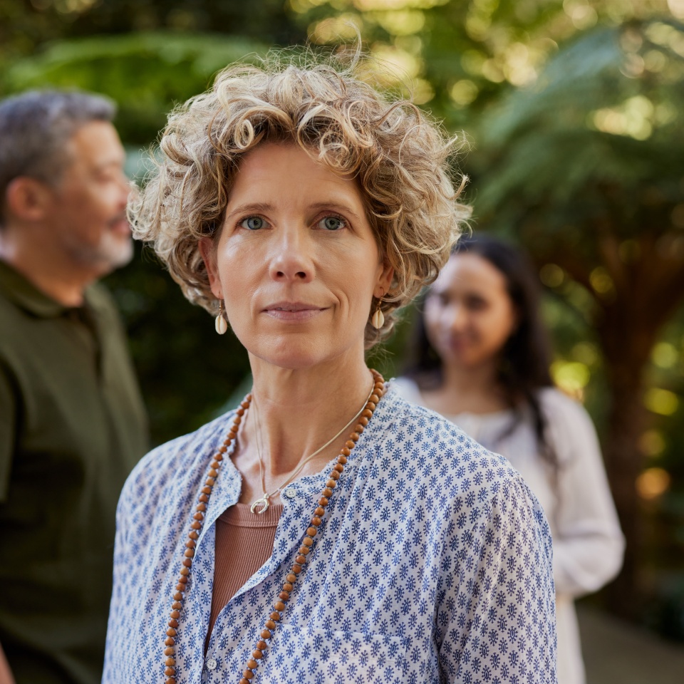 Middle aged woman stands confidently outdoors during a wellness retreat with a group of people behind her. Mid woman in relaxed clothing enjoy a calm moment at a nature based wellness center.