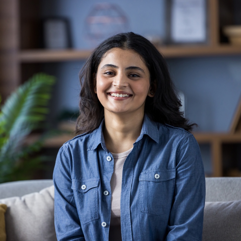 A beautiful young woman sits comfortably at home, smiling radiantly. She is wearing a denim shirt and looks happy and relaxed.