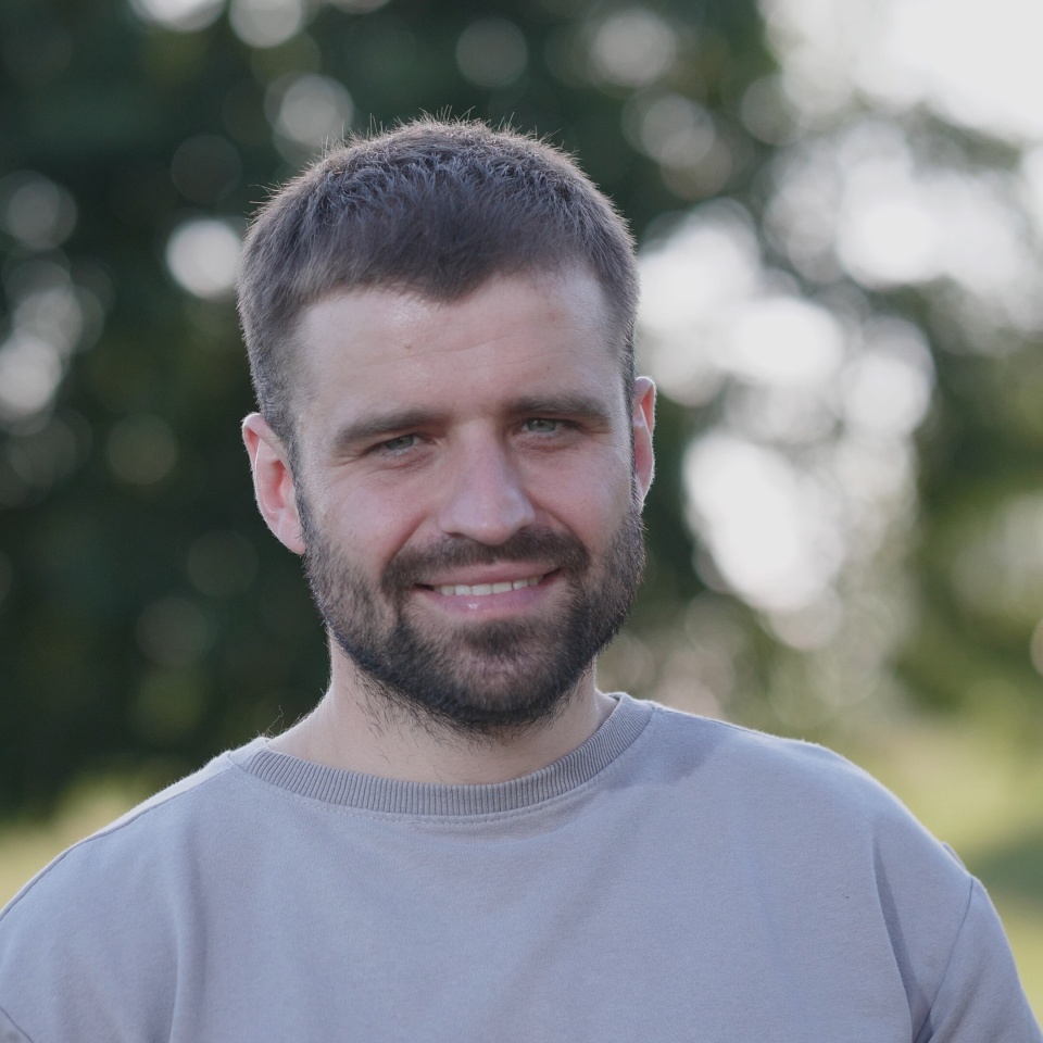 A man with a cheerful expression stands in a park, surrounded by greenery, enjoying a sunny day while exuding a sense of calm and happiness.