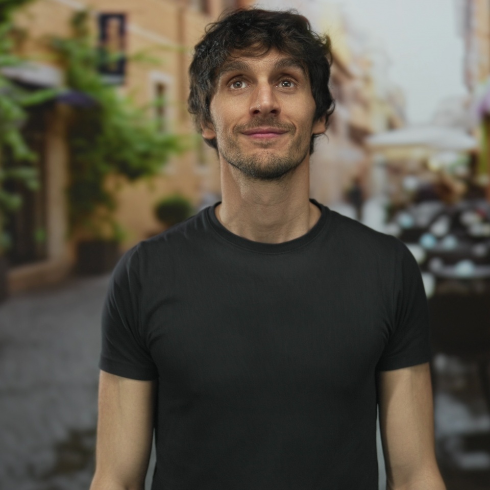 Man in black t shirt smiles while standing on a narrow cobblestone urban street; serenity calm confidence.