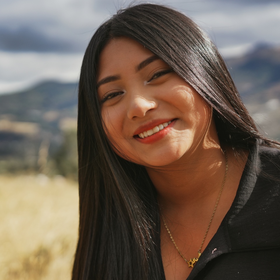 Portrait of a cheerful young latin american woman smiling outdoors with a mountain landscape in the background, enjoying a sunny day in a natural setting