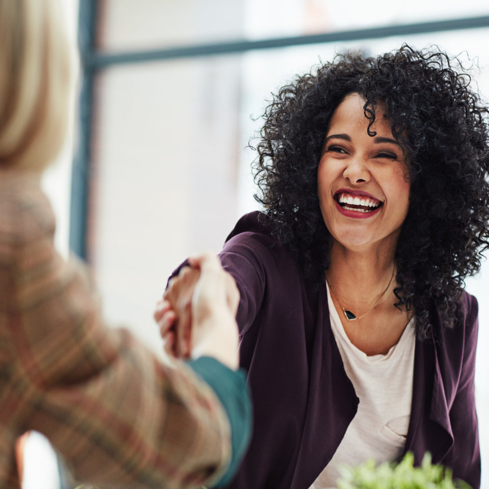 Two women are sitting across from each other in an office setting, smiling and shaking hands.