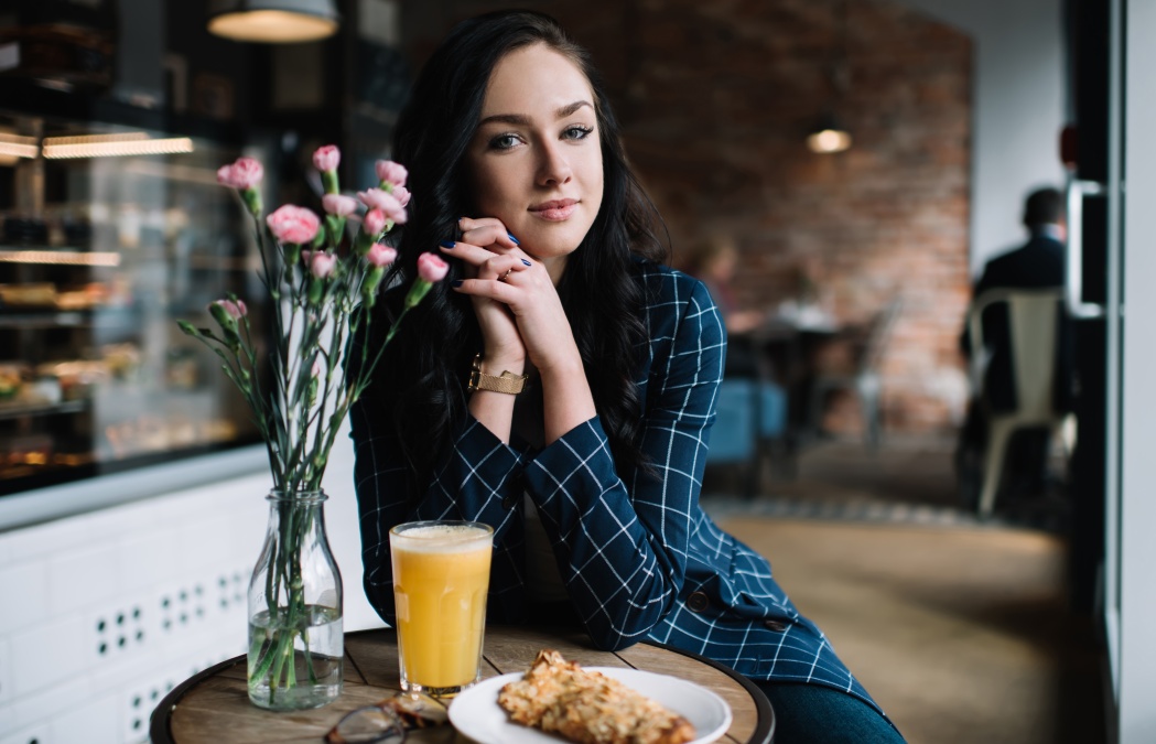 Delighted woman in stylish wear sitting at wooden round table and enjoying fresh juice and cookie during breakfast in cafe